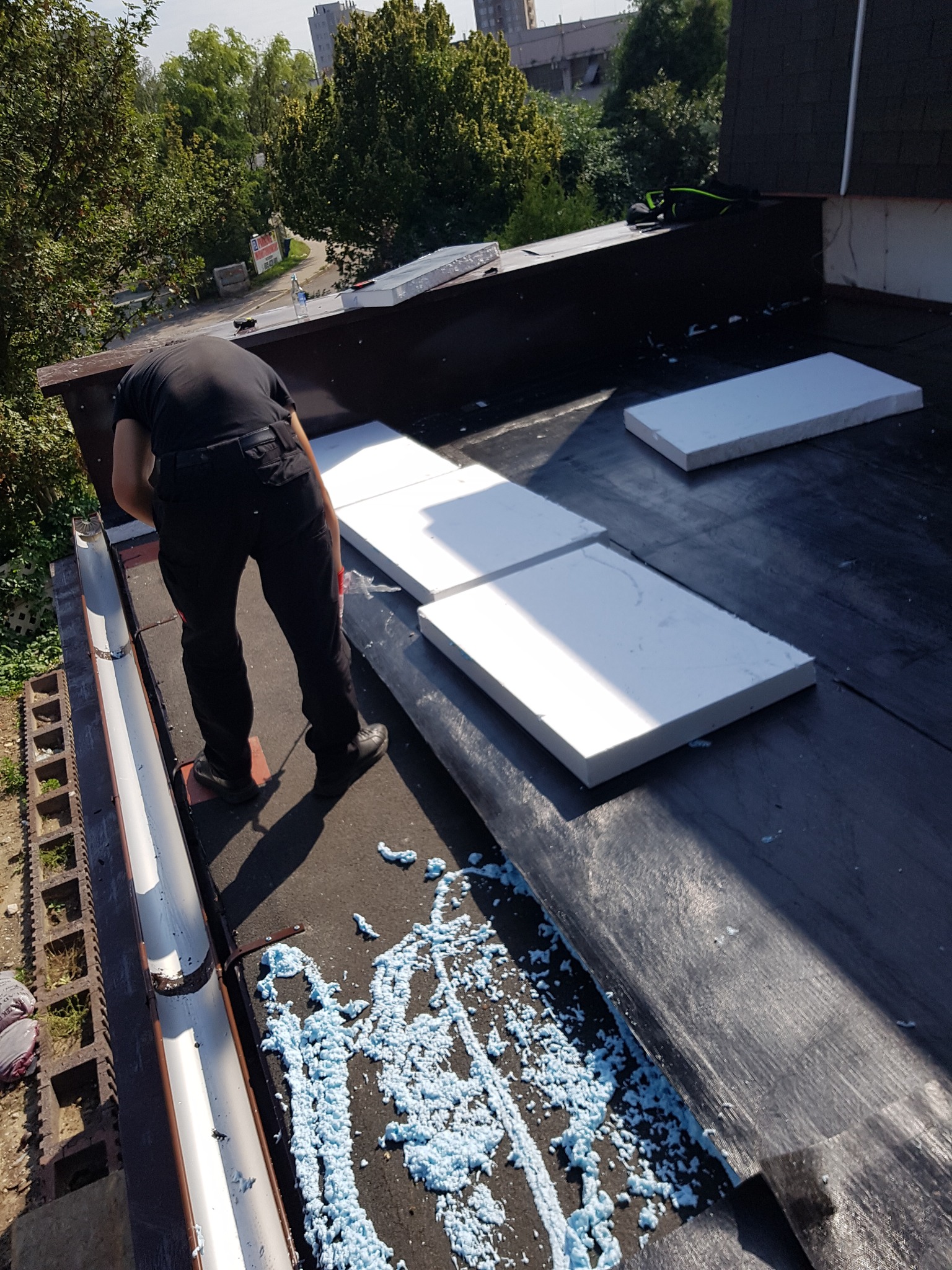 Dekarz w czarnym ubraniu układa białe płyty izolacyjne na czarnym dachu. Widoczne niebieskie pozostałości pianki montażowej i fragment rynny.