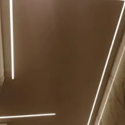 Beżowy sufit z wbudowanym oświetleniem LED w kształcie prostokąta i linii, marmurowe wykończenie ściany w rogu pomieszczenia.