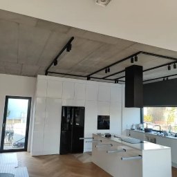 Usługi Remontowo-Budowlane Wykończeniowo Stolarskie Marek Wojak - Nowoczesna kuchnia z białymi szafkami, wyspą kuchenną i betonowym sufitem z czarnym oświetleniem szynowym, widok na zewnątrz przez okno.