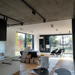 Usługi Remontowo-Budowlane Wykończeniowo Stolarskie Marek Wojak - Wnętrze w trakcie wykańczania: surowy betonowy sufit z czarnymi szynami oświetleniowymi, częściowo umeblowana przestrzeń z widokiem na zewnątrz przez panoramiczne okna, podłoga zabezpieczona folią,...