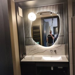 Usługi Remontowo-Budowlane Wykończeniowo Stolarskie Marek Wojak - Nowoczesna łazienka z niesymetrycznym lustrem podświetlanym LED, umywalką nablatową na blacie z imitacji marmuru oraz czarnymi szafkami pod umywalką i dekoracyjnymi pionowymi listwami na ścianie.