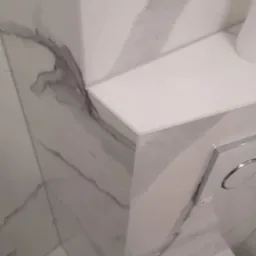 Fragment wykończonej łazienki z marmurowymi płytkami na ścianach i półką nad toaletą ze spłuczką z dwoma przyciskami.