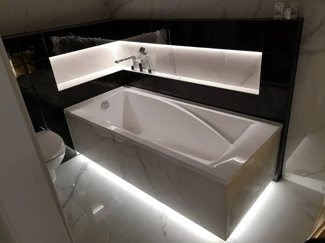 Biała wanna z podświetleniem LED w nowoczesnej łazience z czarnymi płytkami i marmurową podłogą, bateria wannowa na tle wnęki z oświetleniem.
