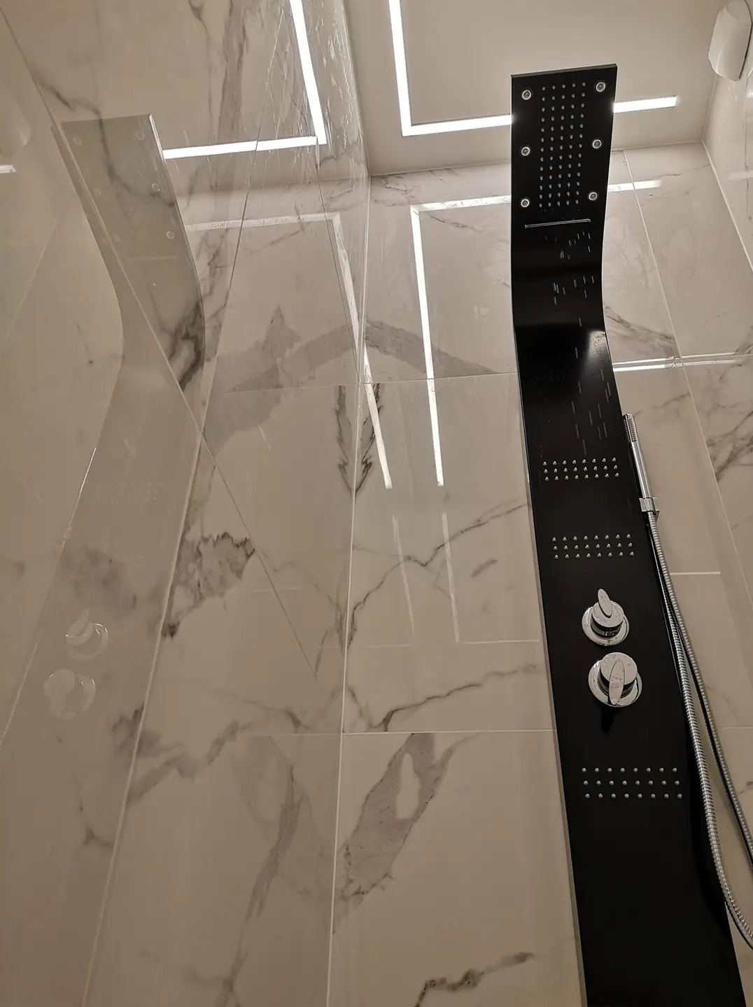 Nowoczesna kabina prysznicowa z czarnym panelem natryskowym na tle marmurowych płytek i oświetlenia LED w suficie.