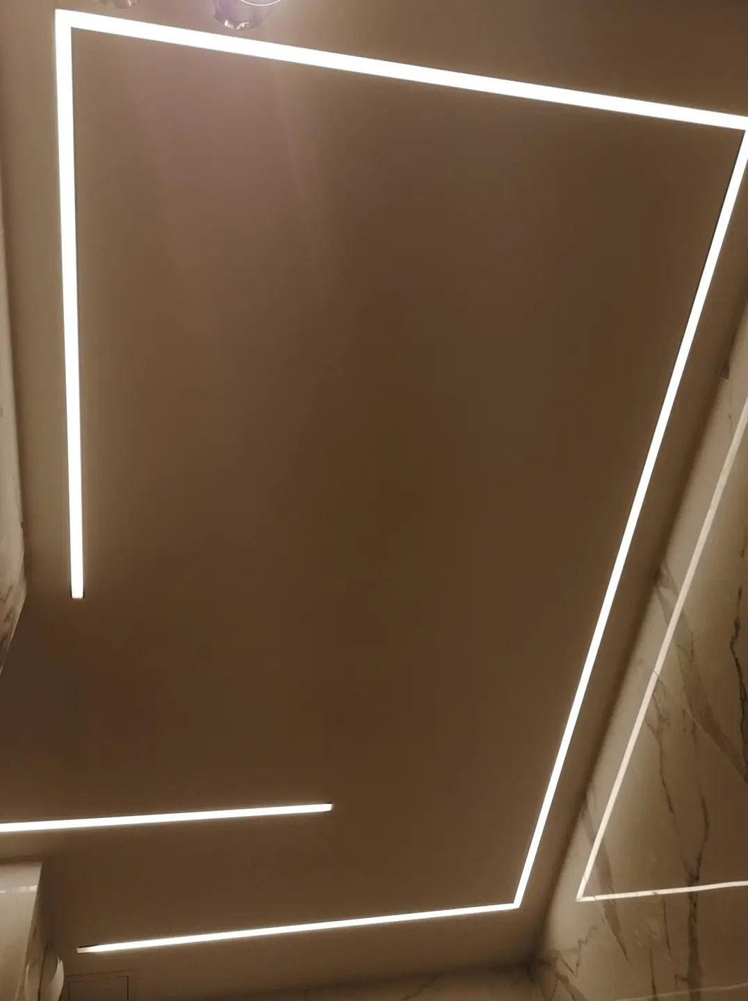 Beżowy sufit z wbudowanym oświetleniem LED w kształcie prostokąta i linii, marmurowe wykończenie ściany w rogu pomieszczenia.