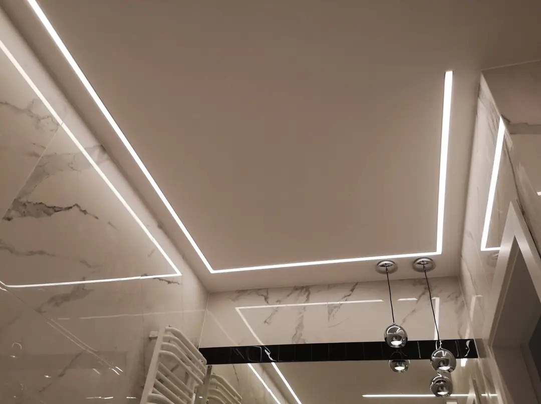 Nowoczesna łazienka w Łęgowie z białymi marmurowymi płytkami i oświetleniem LED w profilach, tworzącym geometryczne wzory na suficie i ścianach, uzupełniona wiszącymi lampami z przezroczystymi...