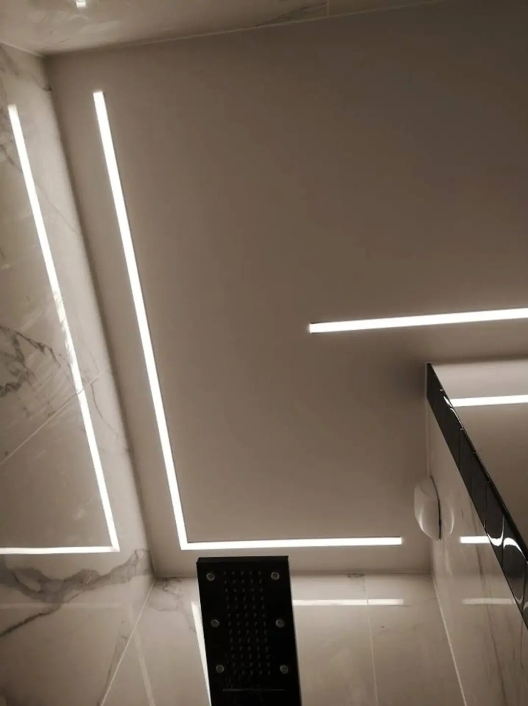 Nowoczesna łazienka z liniowym oświetleniem LED w suficie i na ścianach, wykończona marmurowymi płytkami, z czarną deszczownicą i szklaną kabiną prysznicową.