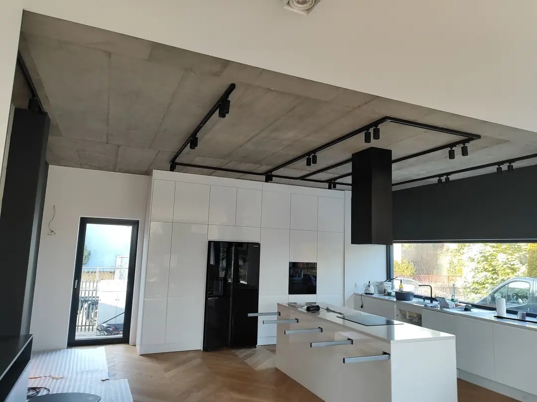 Nowoczesna kuchnia z białymi szafkami, wyspą kuchenną i betonowym sufitem z czarnym oświetleniem szynowym, widok na zewnątrz przez okno.
