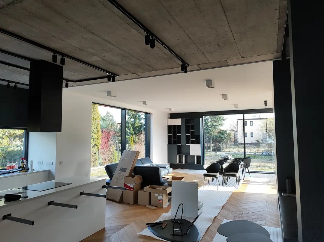 Wnętrze w trakcie wykańczania: surowy betonowy sufit z czarnymi szynami oświetleniowymi, częściowo umeblowana przestrzeń z widokiem na zewnątrz przez panoramiczne okna, podłoga zabezpieczona folią,...