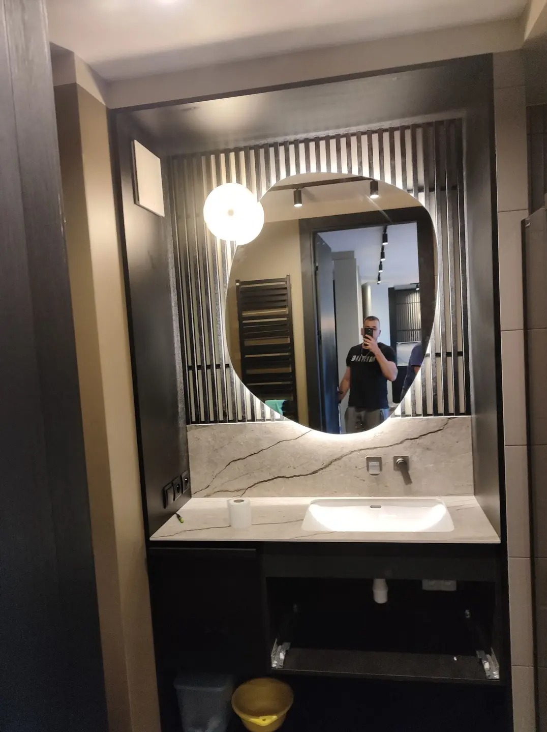 Nowoczesna łazienka z niesymetrycznym lustrem podświetlanym LED, umywalką nablatową na blacie z imitacji marmuru oraz czarnymi szafkami pod umywalką i dekoracyjnymi pionowymi listwami na ścianie.