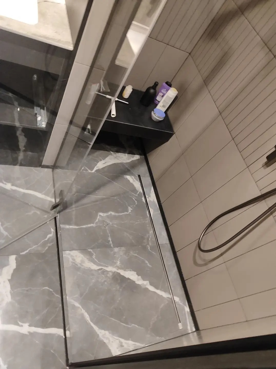 Nowoczesna kabina prysznicowa z szarymi płytkami imitującymi marmur na podłodze i jasnoszarymi płytkami na ścianach, widoczna półka z kosmetykami i zestawem do czyszczenia, ujęcie z góry.