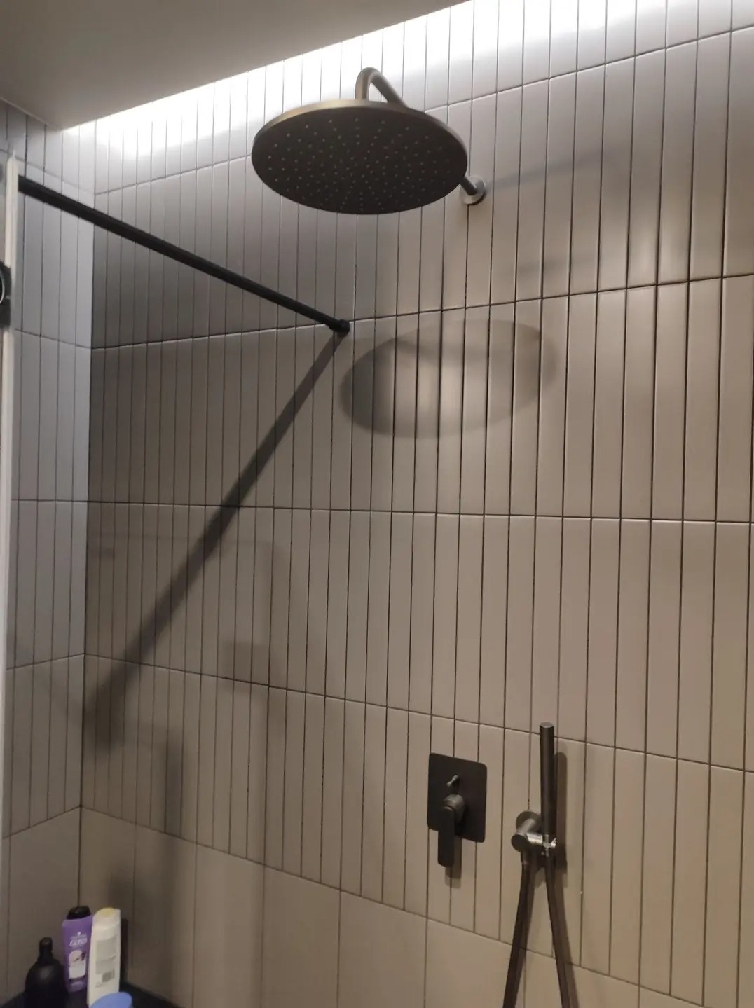 Nowoczesna kabina prysznicowa z deszczownicą w kolorze grafitowym, minimalistyczna armatura, podświetlenie LED wzdłuż sufitu, wąskie płytki w odcieniach szarości.