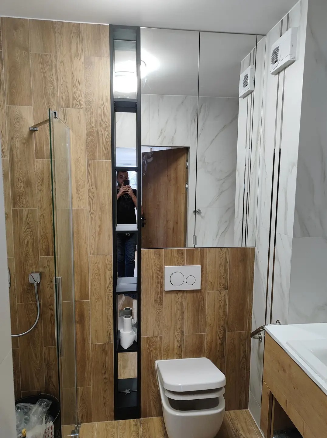 Wykończona łazienka z drewnopodobnymi płytkami, kabiną prysznicową ze szklanymi drzwiami, zabudową lustrzaną z czarną wnęką na akcesoria i toaletą wiszącą z przyciskiem dwudzielnym.