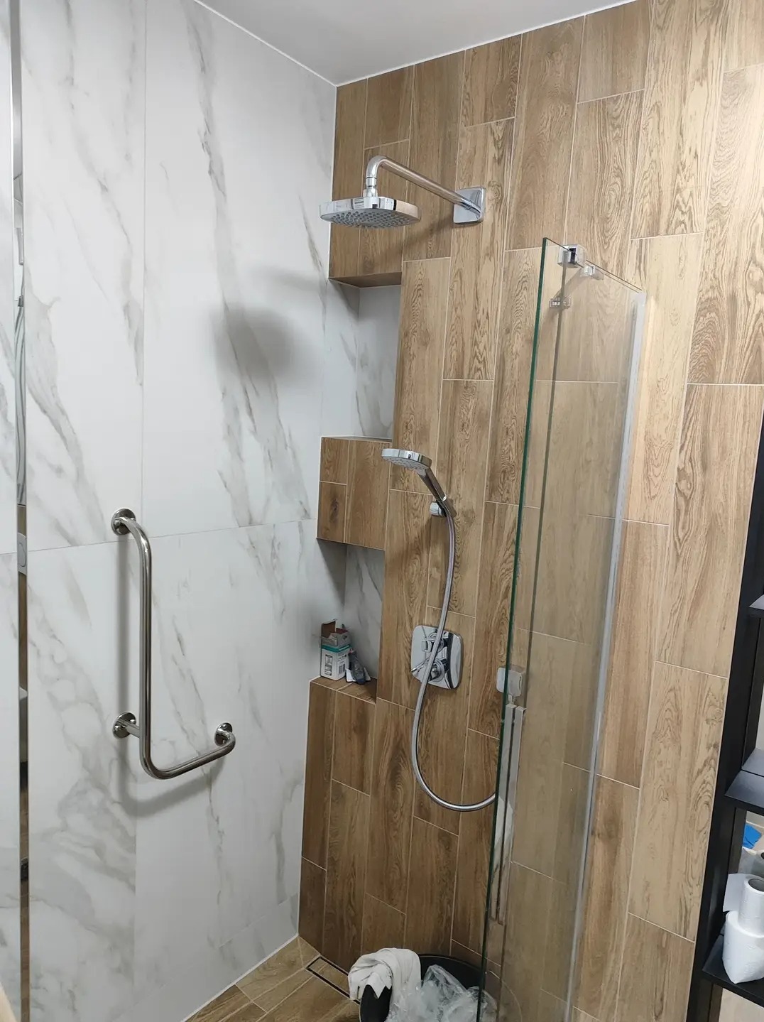 Nowoczesna kabina prysznicowa z deszczownicą i uchwytem dla niepełnosprawnych, wykończona płytkami imitującymi drewno i marmur, z wnęką na kosmetyki.