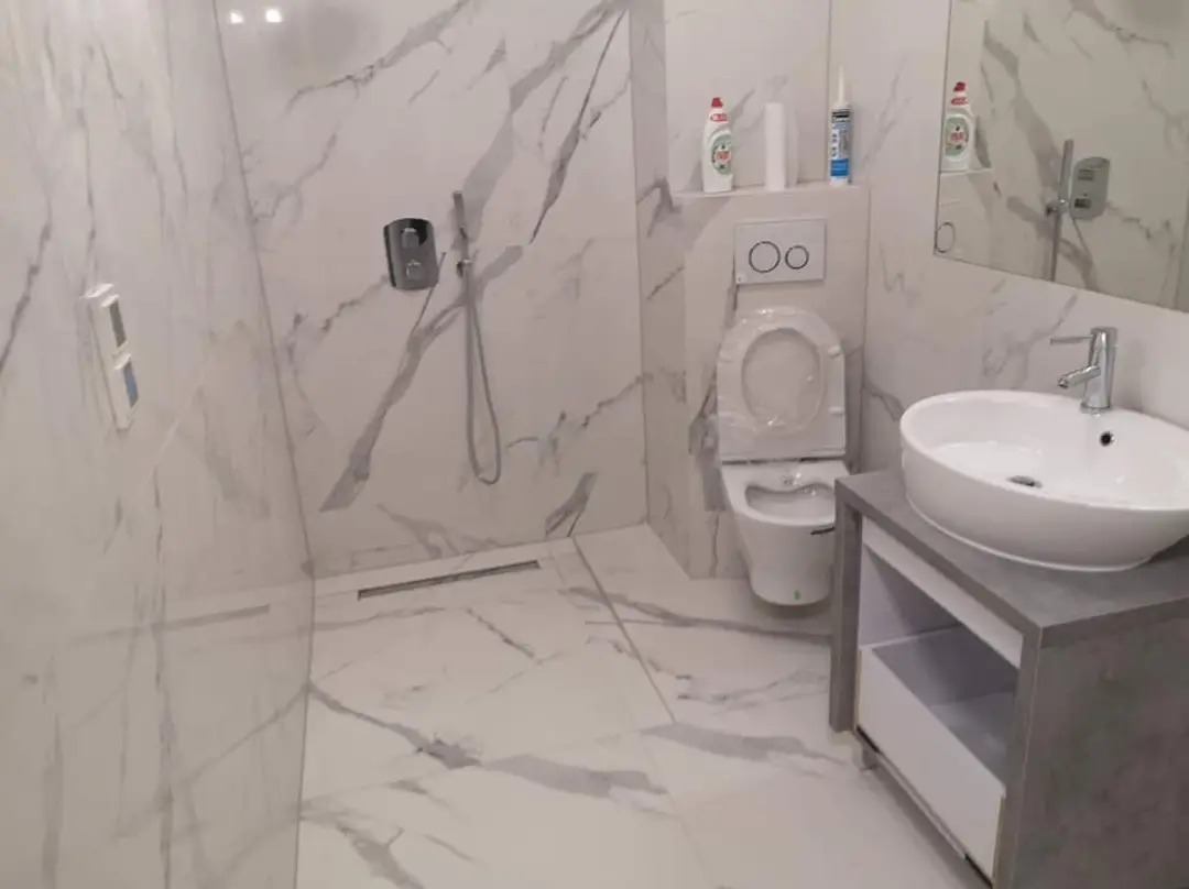 Nowoczesna łazienka z białymi marmurkowymi płytkami, wiszącą toaletą z folią ochronną, umywalką nablatową na szarej szafce i odpływem liniowym w podłodze.