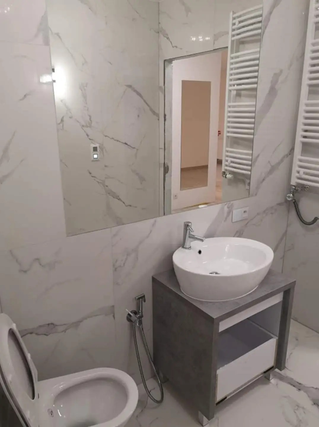 Nowoczesna łazienka z marmurowymi płytkami, umywalką nablatową na szarej szafce, wiszącym grzejnikiem drabinkowym i bidetką przy toalecie.