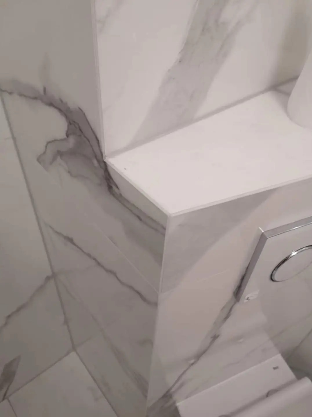 Fragment wykończonej łazienki z marmurowymi płytkami na ścianach i półką nad toaletą ze spłuczką z dwoma przyciskami.