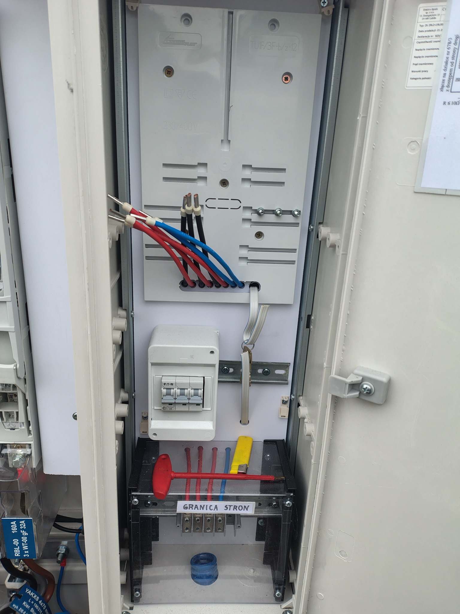 Otwarta skrzynka elektryczna z widocznymi przewodami (czerwone, niebieskie, czarne), bezpiecznikami Eaton, listwą zaciskową z oznaczeniem 'Granica Stron' oraz elementami montażowymi.