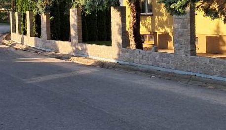 Nowoczesne ogrodzenie modułowe z betonowych bloczków w kolorze piaskowym, oddzielające posesję od ulicy, z widocznymi słupkami i przęsłami, na tle żółtego budynku i zielonych drzew.