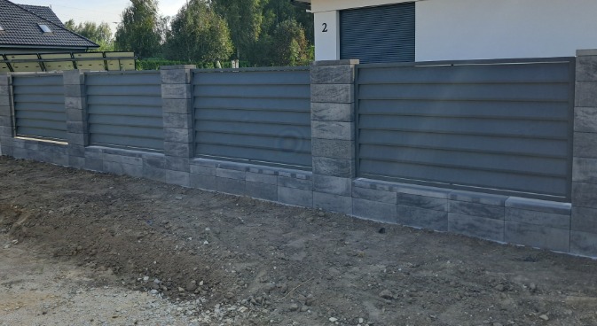 Nowoczesne ogrodzenie panelowe w kolorze szarym z kamiennymi słupkami i podmurówką, widoczne na tle domu z numerem 2 i zielenią.