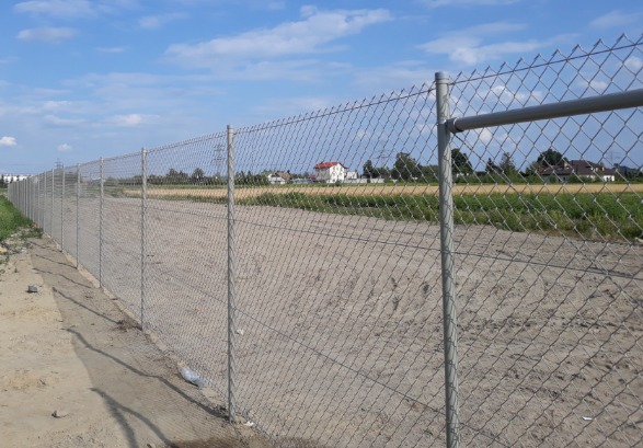 Długa, szara siatka ogrodzeniowa rozciągająca się wzdłuż piaszczystego terenu, z widocznymi domami w tle pod błękitnym niebem.
