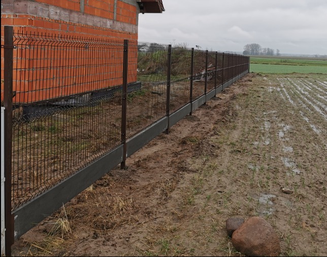 Nowoczesne ogrodzenie panelowe z podmurówką betonową, widok z boku na tle pola i budynku z czerwonej cegły w pochmurny dzień.