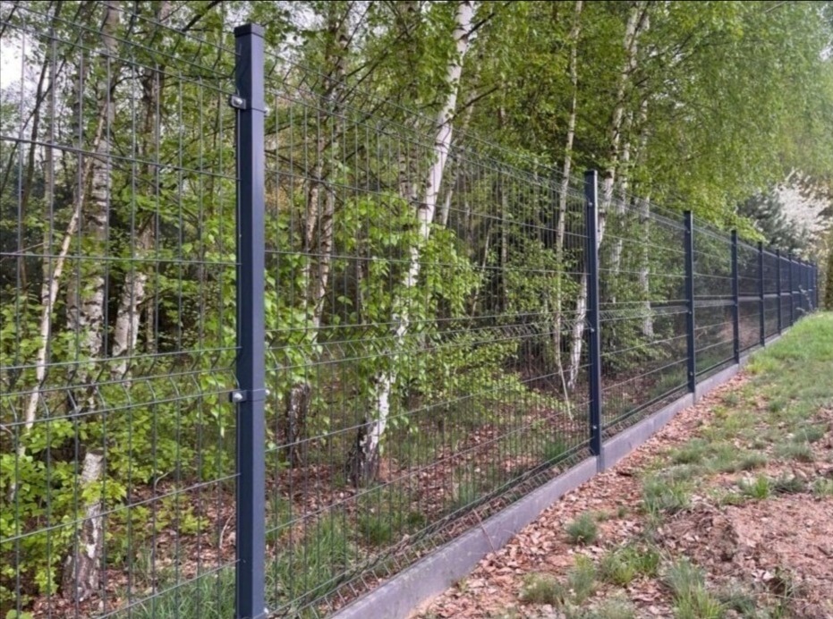Długi odcinek panelowego ogrodzenia z ciemnej siatki, z betonową podmurówką, biegnący wzdłuż granicy działki z widocznymi drzewami w tle.