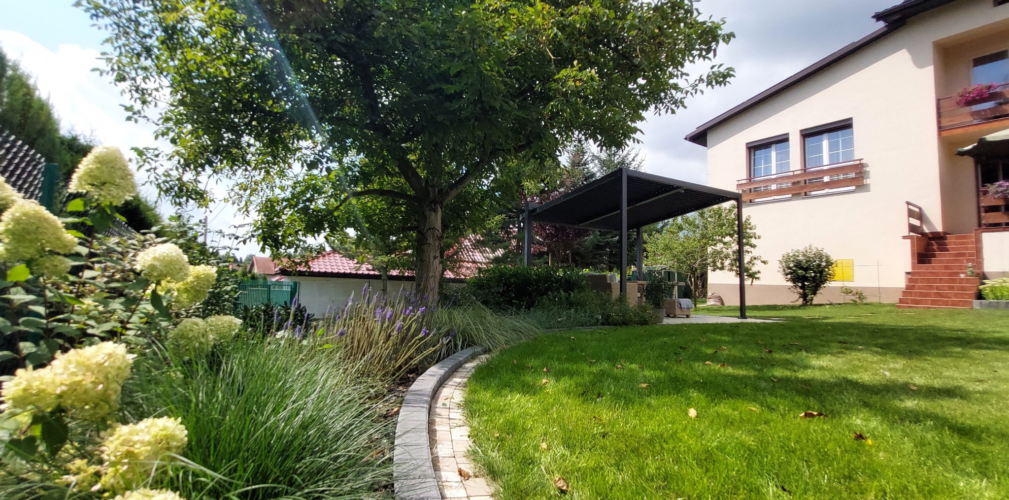 Ogród przydomowy z pergolą, ścieżką z kostki brukowej, trawą i kwitnącymi hortensjami.