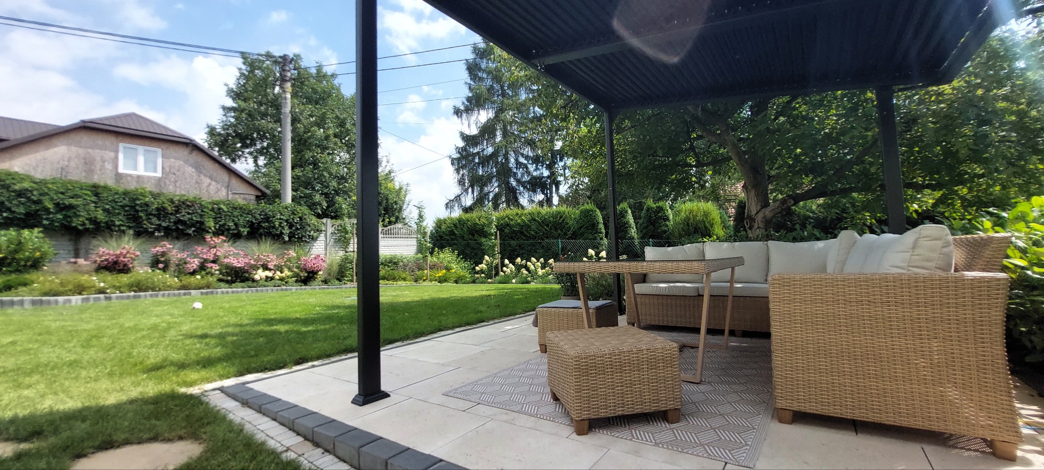 Zadaszony taras z meblami ogrodowymi z rattanu, widok na zadbany trawnik, rabaty kwiatowe z hortensjami i strzyżone żywopłoty w tle.