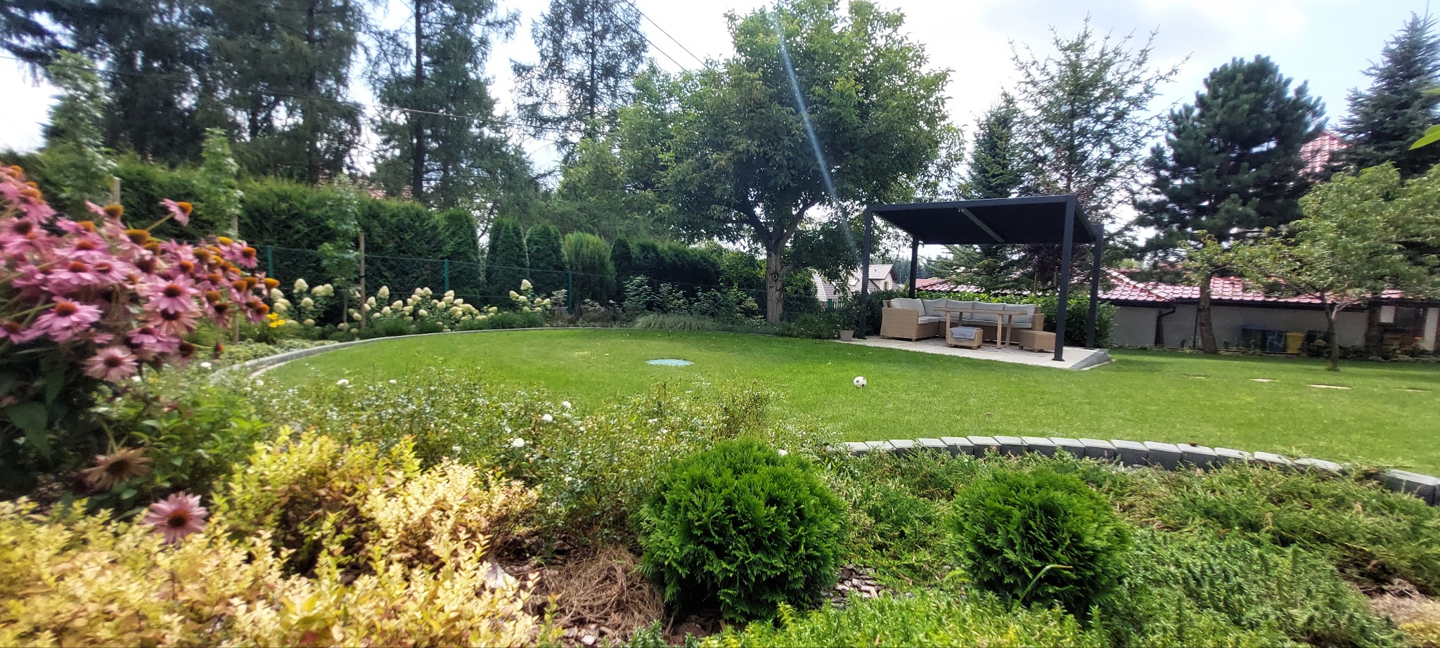 Pełen zieleni ogród z równo przystrzyżonym trawnikiem, rabatami kwiatowymi i krzewami, zadaszoną strefą relaksu z meblami ogrodowymi w tle.