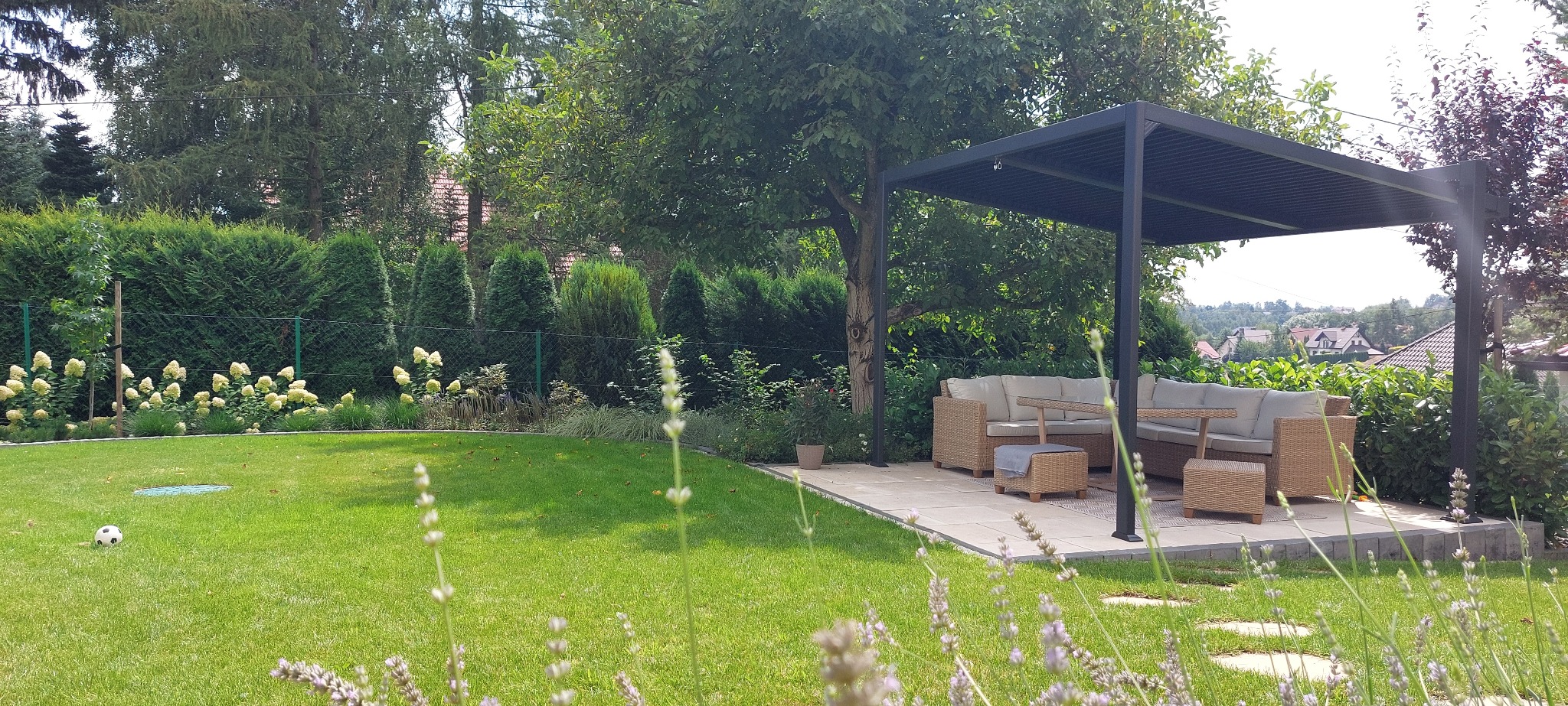 Nowoczesna pergola z rattanowym zestawem wypoczynkowym na kamiennej platformie, otoczona bujną zielenią trawnika i rabatami kwiatowymi, z piłką do gry leżącą na trawie.