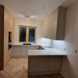 Jasna kuchnia z drewnianą podłogą ułożoną w jodełkę, okno zabezpieczone folią, oświetlenie podszafkowe