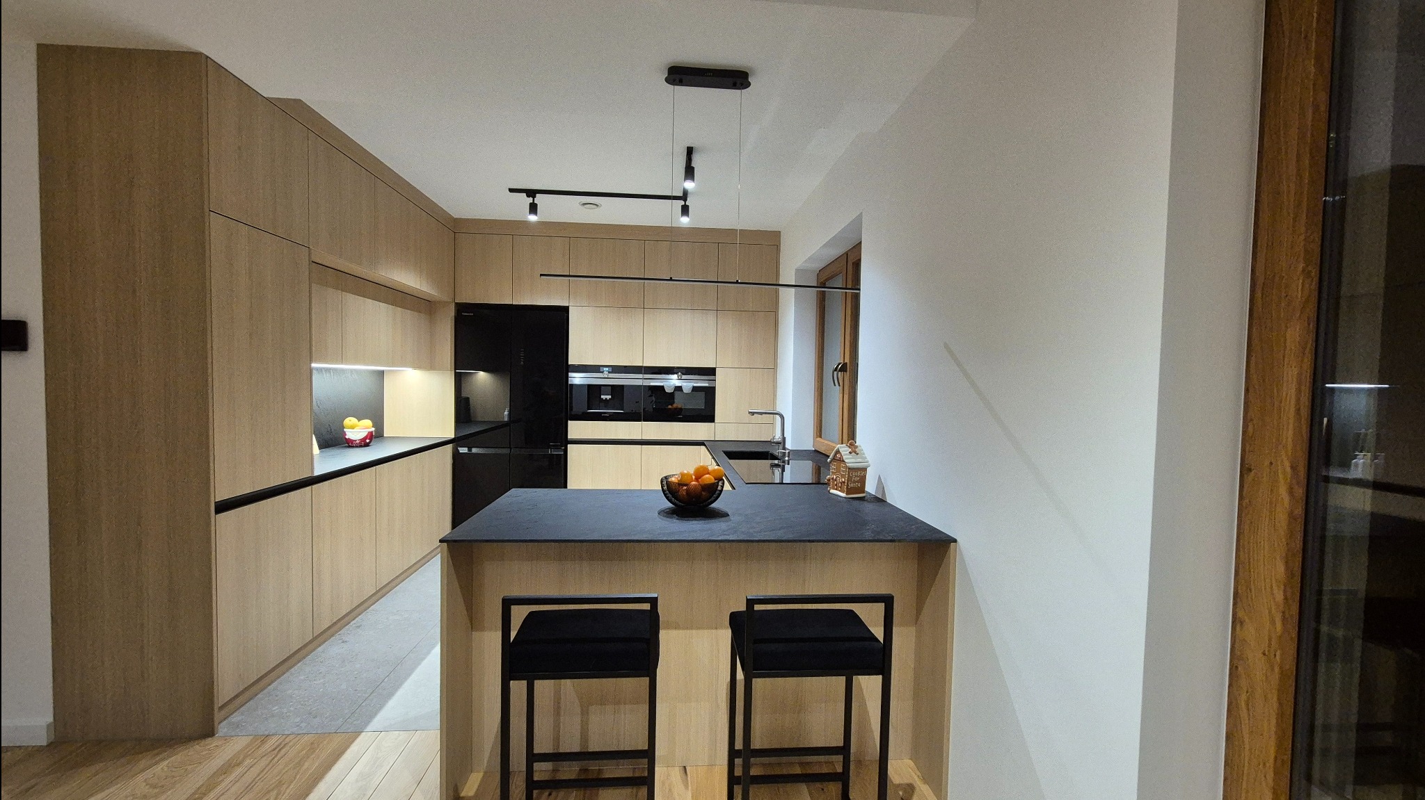 Nowoczesna kuchnia z drewnianymi frontami, czarnym blatem i wyspą z dwoma czarnymi hokerami, oświetlona listwą LED i lampami wiszącymi