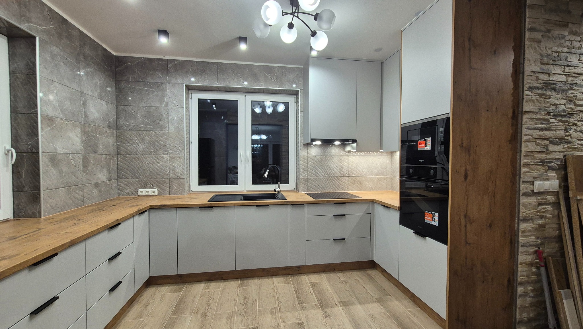 Jasnoszara kuchnia w kształcie litery U z drewnianymi blatami, czarnym zlewem i płytą indukcyjną, oświetlona nowoczesną lampą sufitową i podświetleniem szafek, na ścianie dekoracyjne płytki...