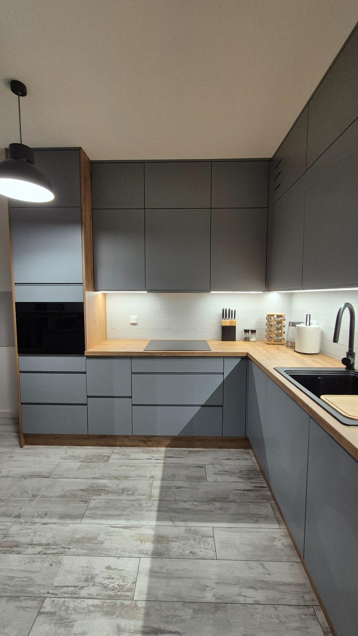 Nowoczesna kuchnia w odcieniach szarości z drewnianymi akcentami, oświetleniem LED podszafkowym i płytkami imitującymi cegłę na ścianie, z widocznymi akcesoriami kuchennymi.