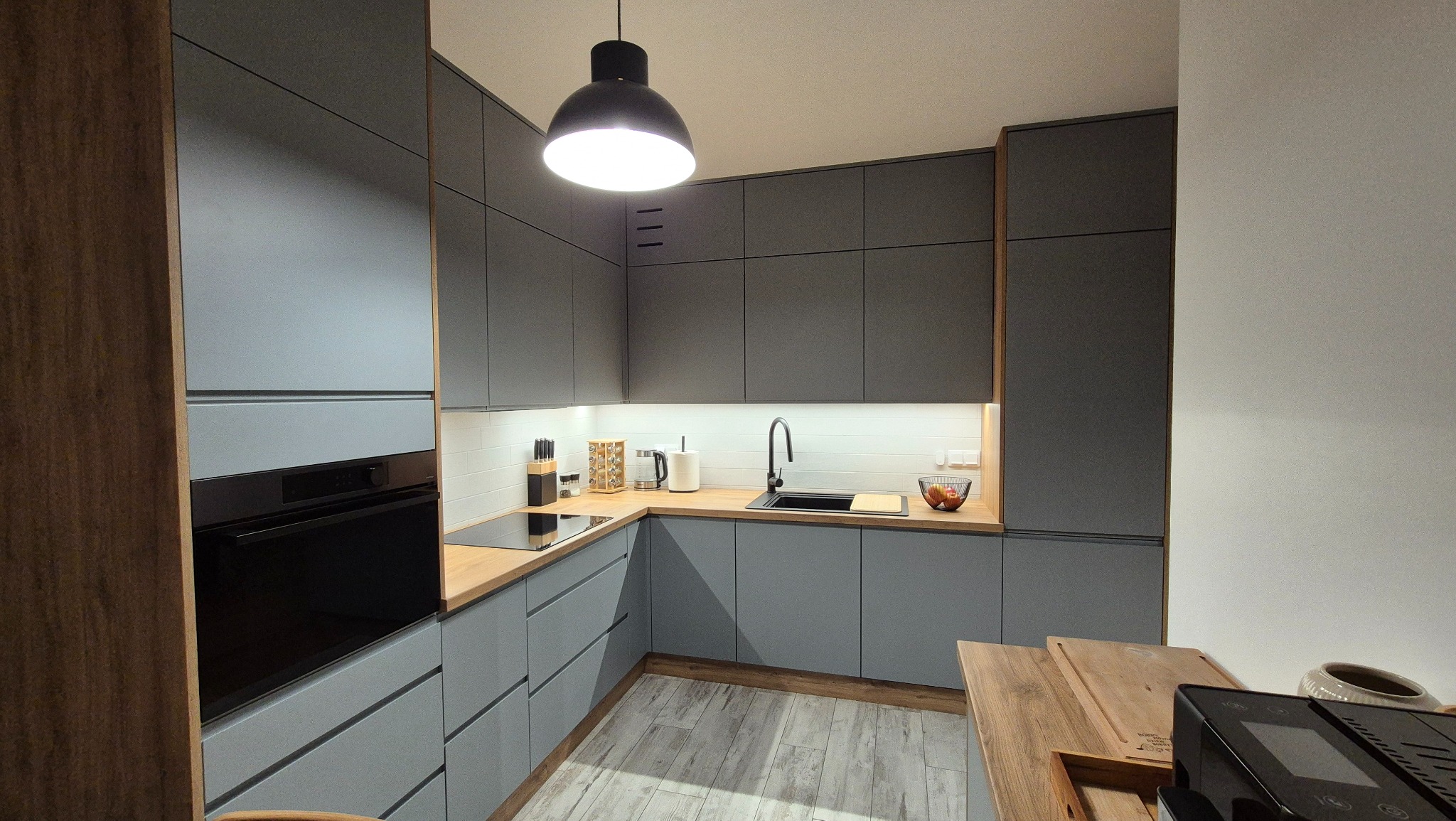 Szara kuchnia z drewnianymi akcentami, czarnym zlewem i płytą indukcyjną podświetlona lampą sufitową, widok na blat z akcesoriami kuchennymi i owocami w misce.