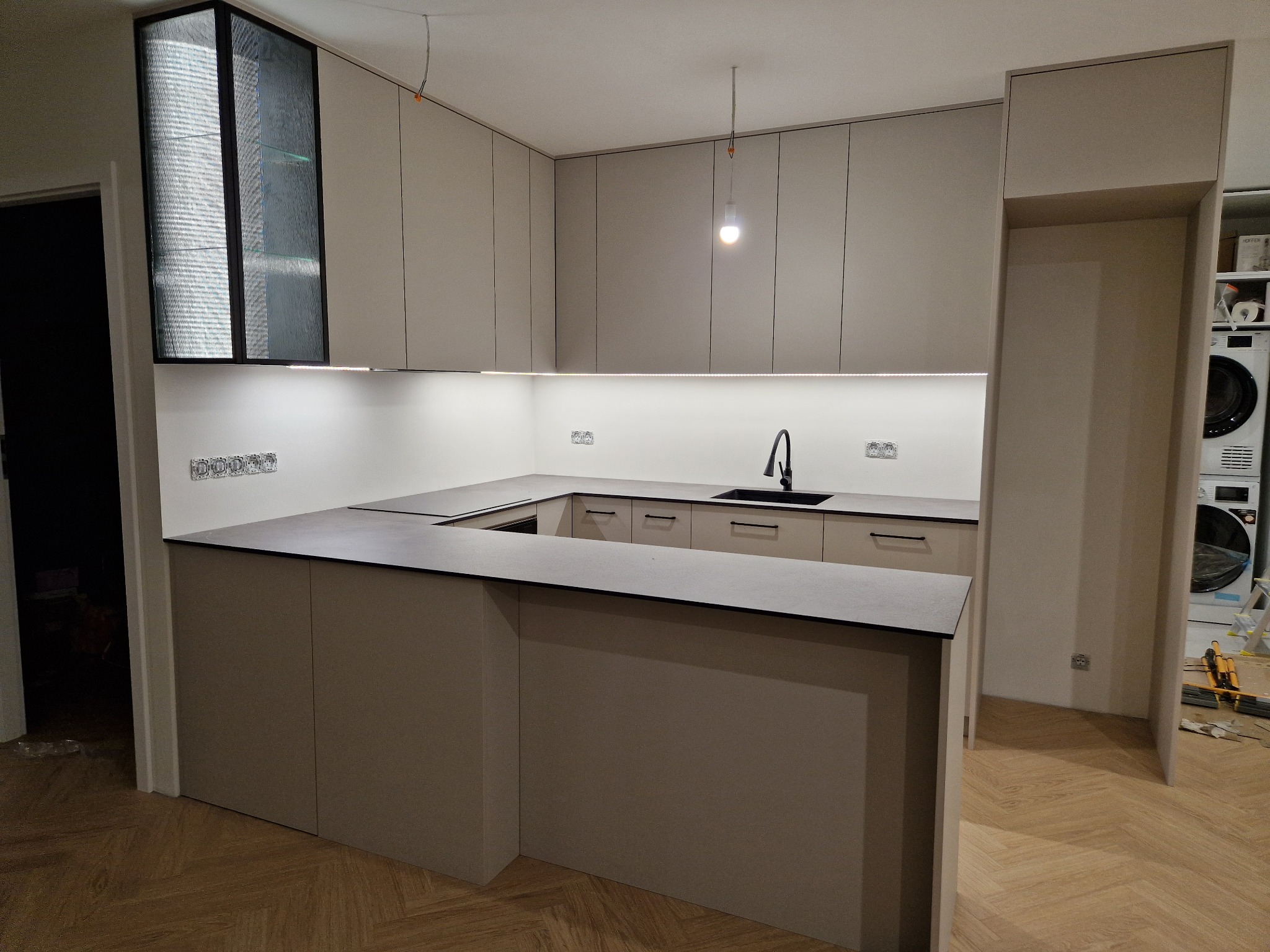 Nowoczesna kuchnia z wyspą, fronty w kolorze beżowym, czarny blat i bateria, ryflowana szyba w górnej szafce, parkiet w jodełkę, widoczne gniazdka i lampa bez klosza.