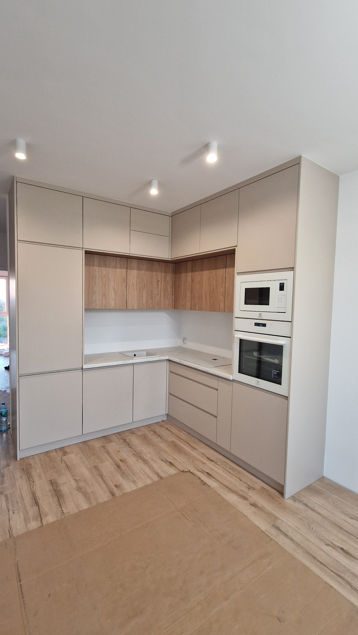 Jasna kuchnia z drewnianymi akcentami, zabudowana pod sufit, z wbudowanym piekarnikiem i mikrofalówką, zabezpieczona kartonem na podłodze podczas montażu.