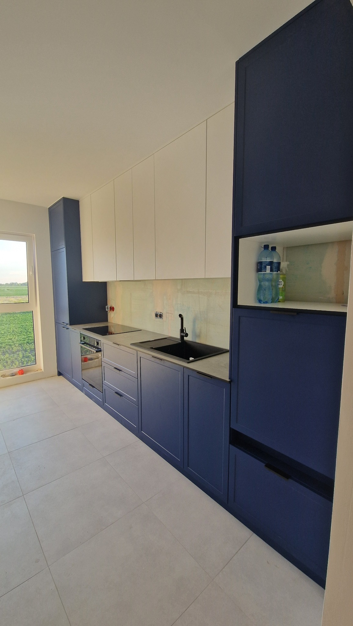 Nowoczesna kuchnia z granatowymi dolnymi szafkami i białymi górnymi, z czarnym zlewem i płytą grzewczą, widok na zielone pole przez okno.