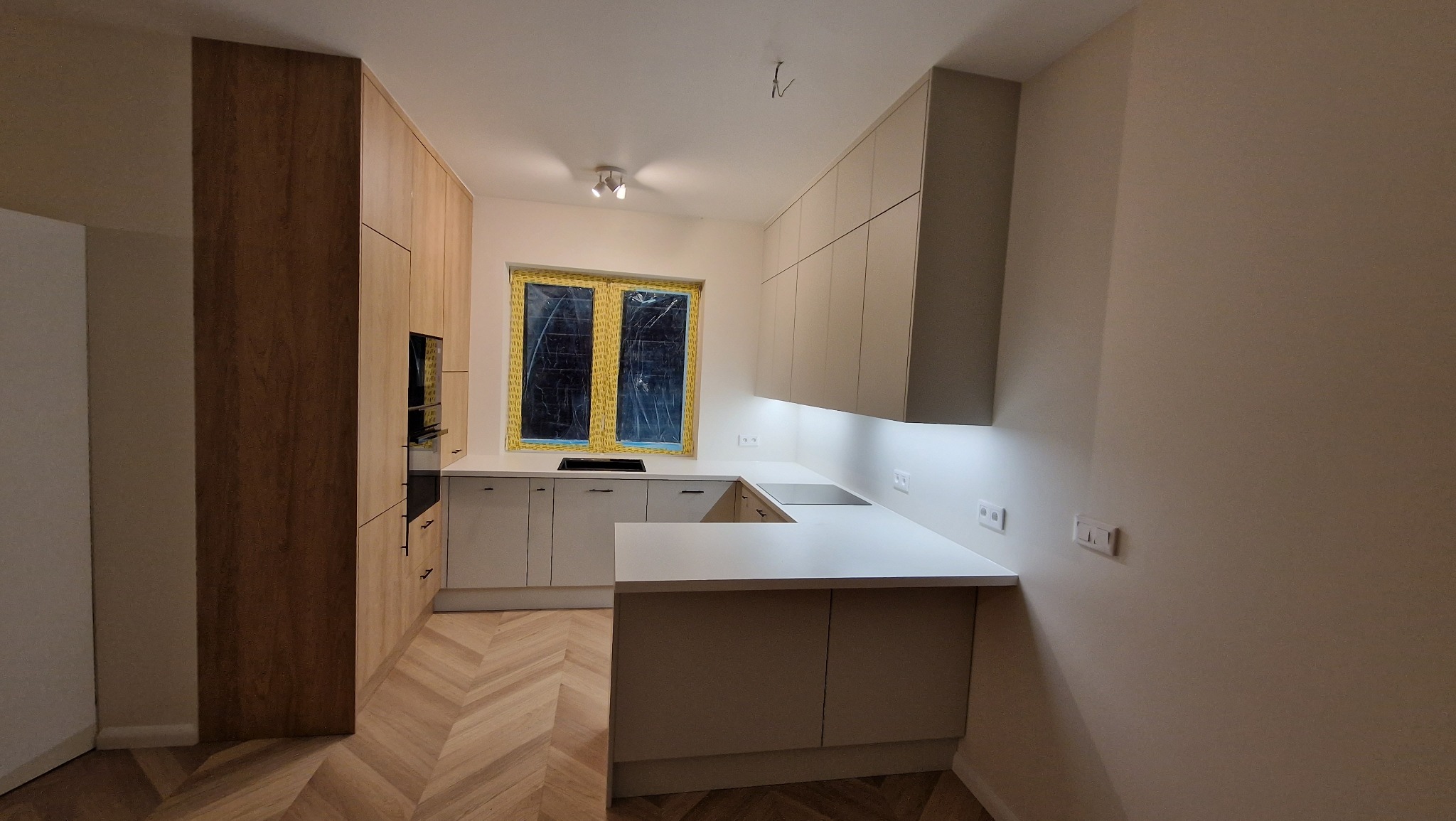 Jasna kuchnia z drewnianą podłogą ułożoną w jodełkę, okno zabezpieczone folią, oświetlenie podszafkowe