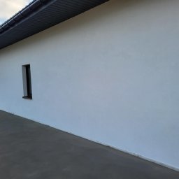 DARIUSZ BORÓWKA DAREX - Boczny widok domu z białą elewacją i ciemnym dachem. Widoczne małe okno i betonowy chodnik. Minimalistyczny styl, równomierne oświetlenie.