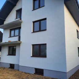 DARIUSZ BORÓWKA DAREX - Biała elewacja dwukondygnacyjnego domu z ciemnymi oknami i szarą podmurówką, widok z boku, częściowo widoczne rusztowanie balkonu, błękitne niebo w tle.