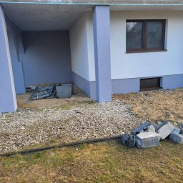 DARIUSZ BORÓWKA DAREX - Świeżo otynkowana elewacja domu jednorodzinnego z szarą podmurówką i kolumnami. Na pierwszym planie gruz i kamienie, sugerujące prace wykończeniowe.