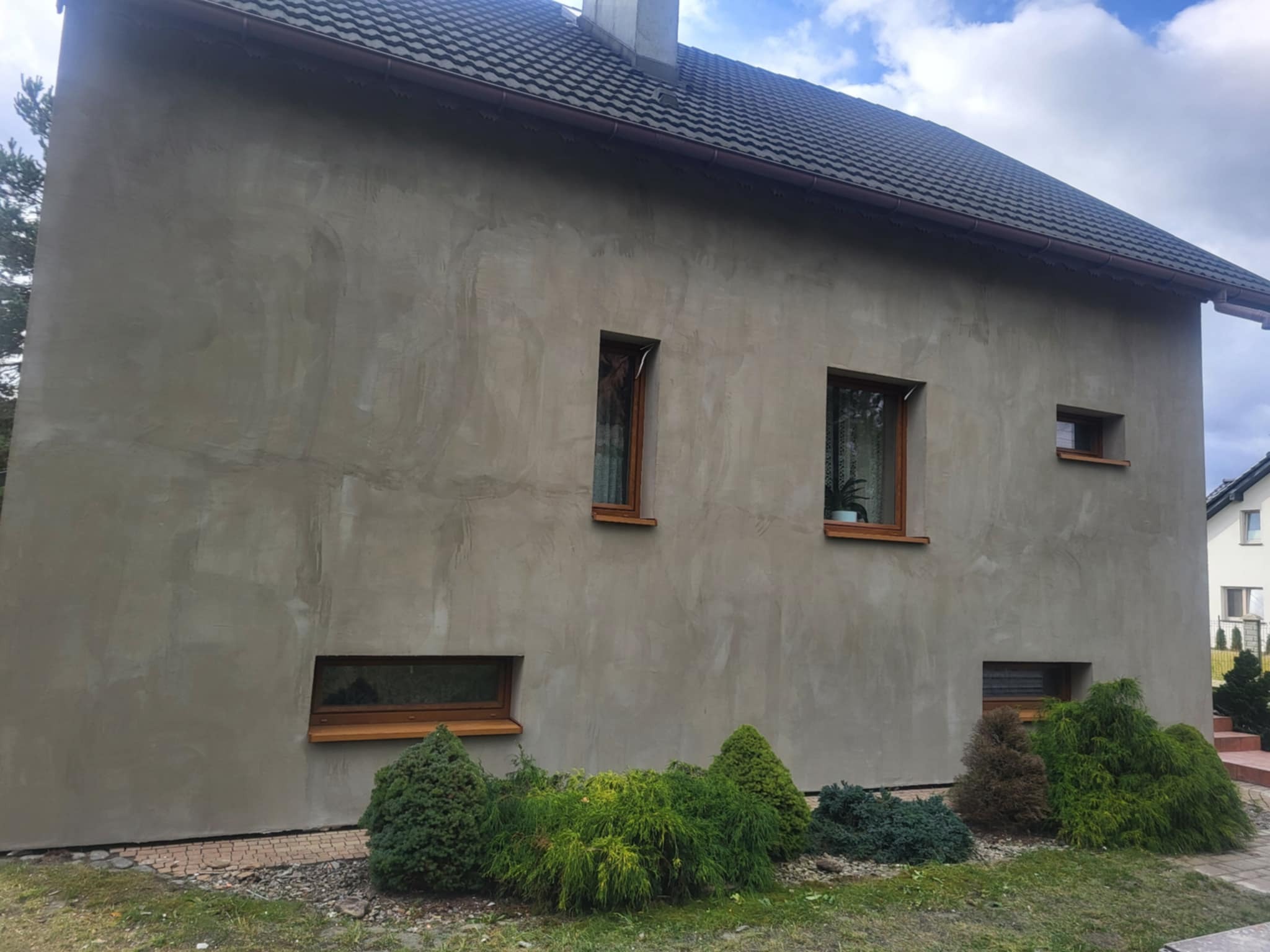 Surowa elewacja domu jednorodzinnego z drewnianą stolarką okienną, widoczna faktura tynku przed malowaniem, zieleń w ogrodzie. Ujęcie z boku budynku.