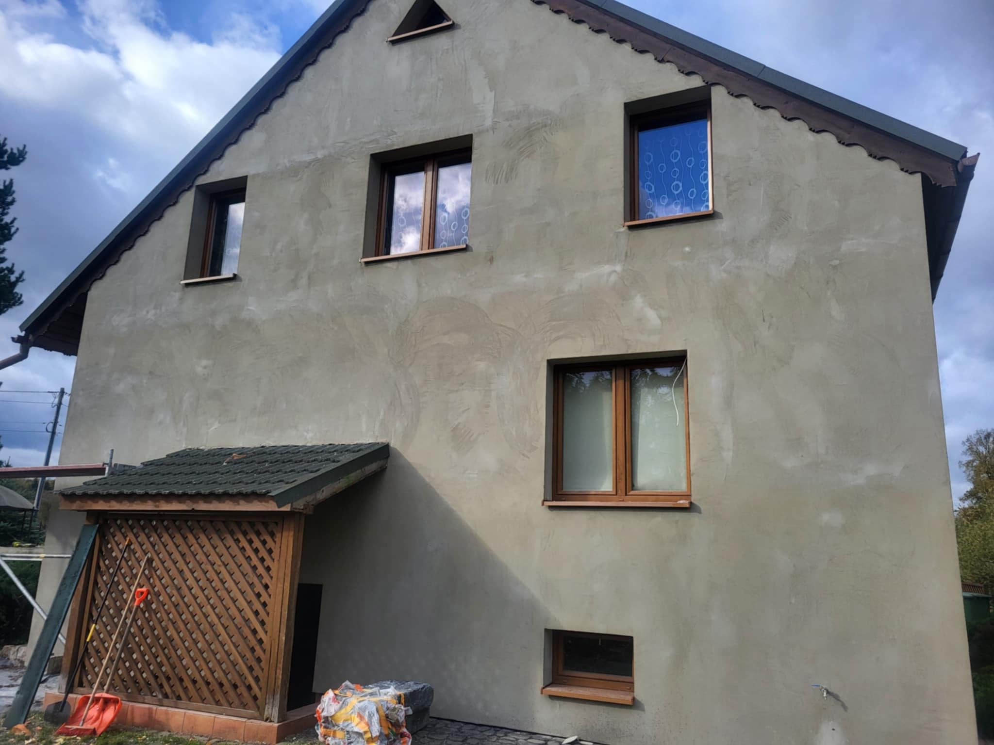 Dom jednorodzinny z szarą elewacją w trakcie prac wykończeniowych, widoczne ślady po nakładaniu tynku. Na pierwszym planie narzędzia budowlane i materiały.