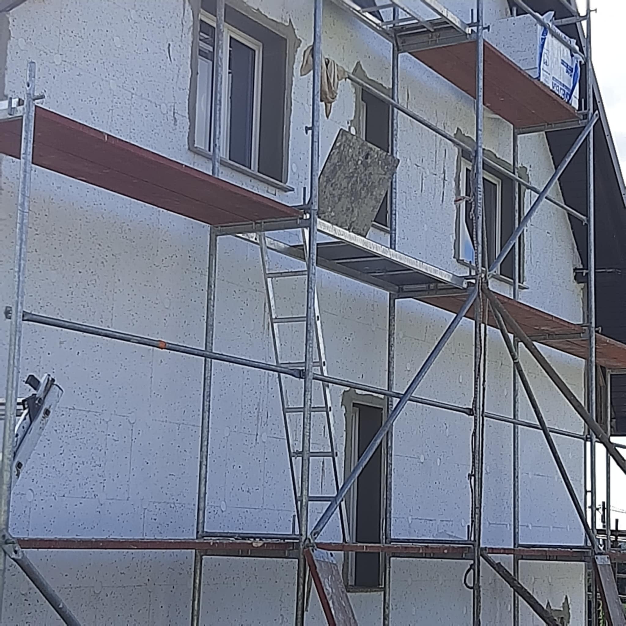 Dom w trakcie ocieplania, widoczne rusztowania i białe płyty izolacyjne na elewacji. Częściowo zakończone prace, widoczne okna i fragment dachu.