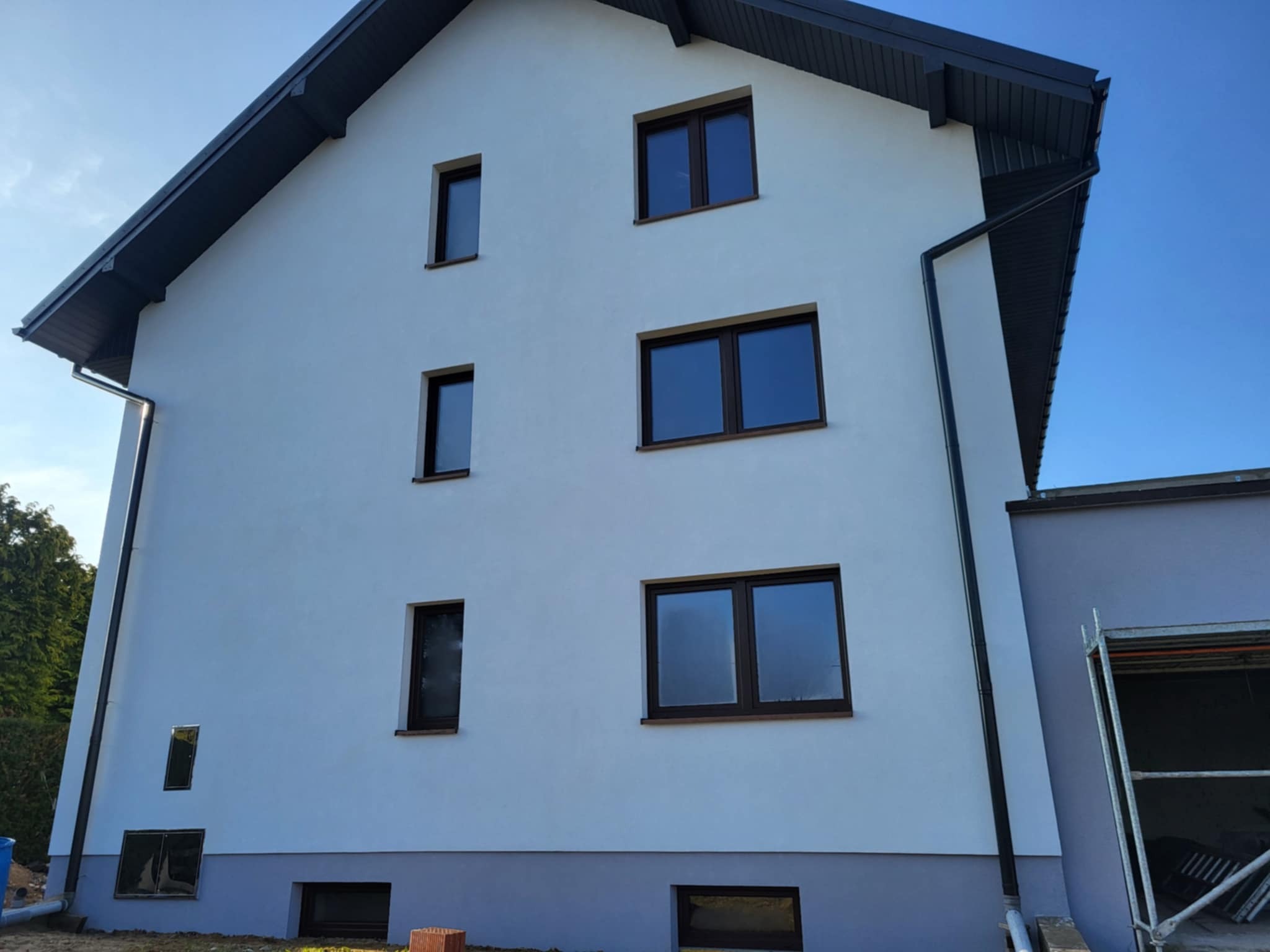 Biała elewacja domu jednorodzinnego z ciemnymi oknami i dachem, widok z dołu, fragment rusztowania z prawej strony. Nowoczesny wygląd.