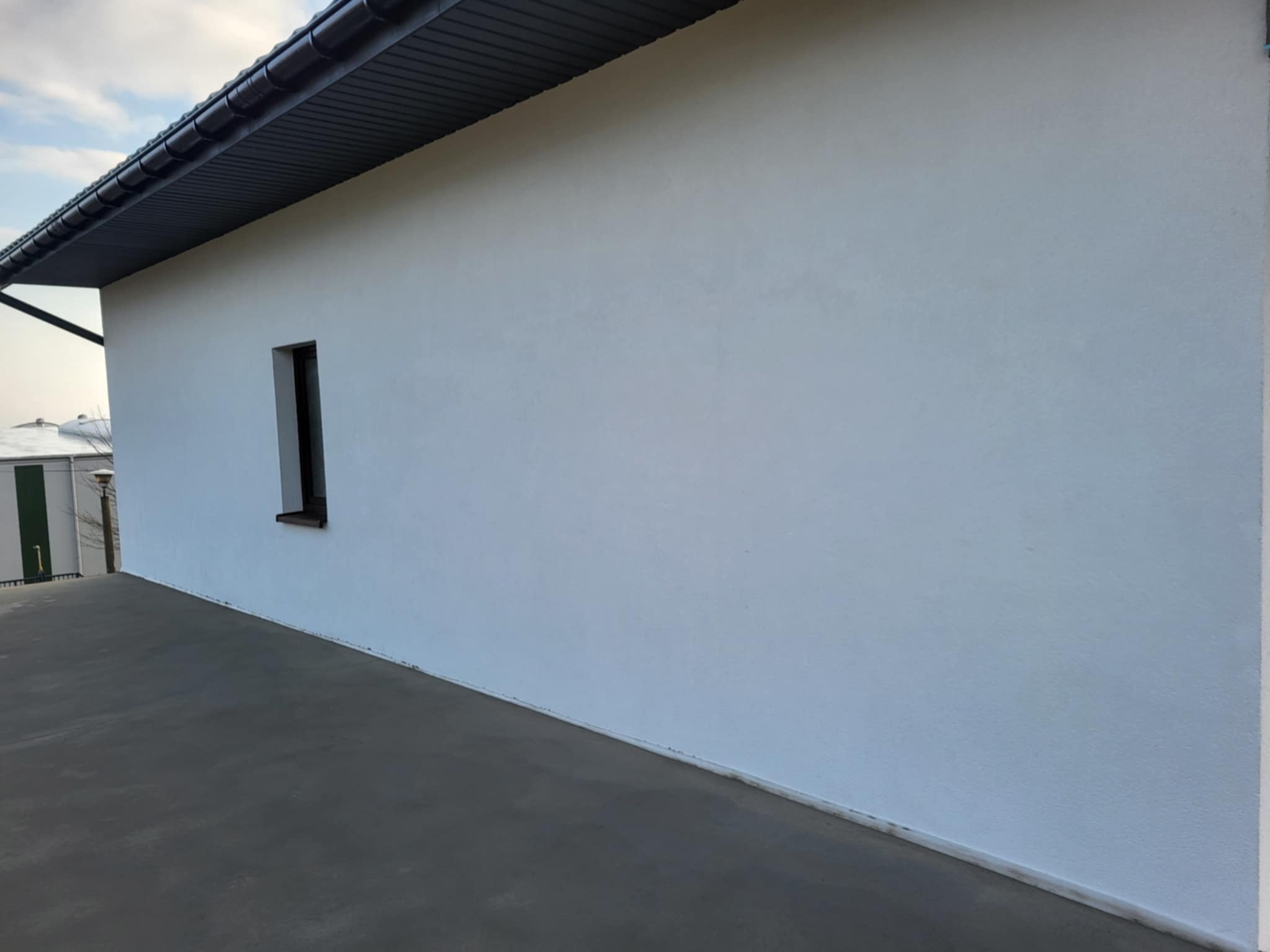 Boczny widok domu z białą elewacją i ciemnym dachem. Widoczne małe okno i betonowy chodnik. Minimalistyczny styl, równomierne oświetlenie.