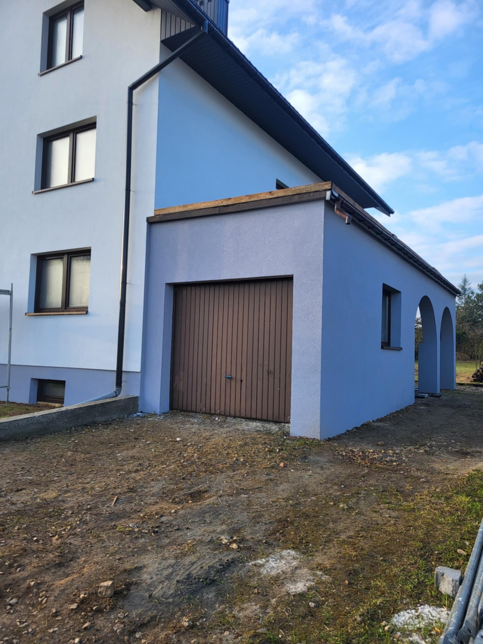 Dwukolorowa elewacja domu z garażem: biała część główna i niebieska dobudówka z brązową bramą garażową. Widoczne okna i fragment terenu.