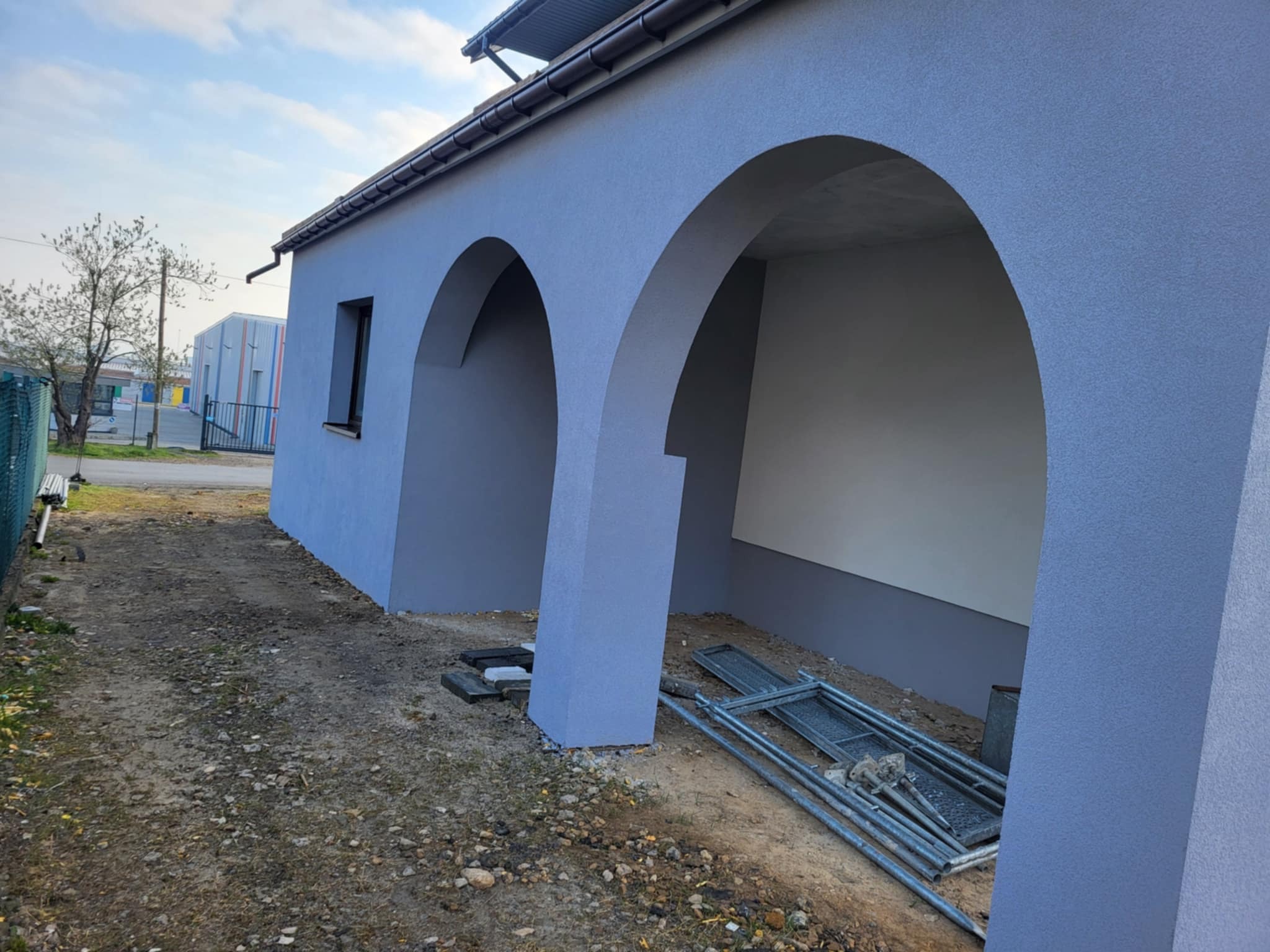 Niebieska elewacja budynku z łukami, widok z boku. Na ziemi leżą elementy rusztowania. Świeżo pomalowana ściana z widoczną fakturą. Budynek w trakcie prac wykończeniowych.